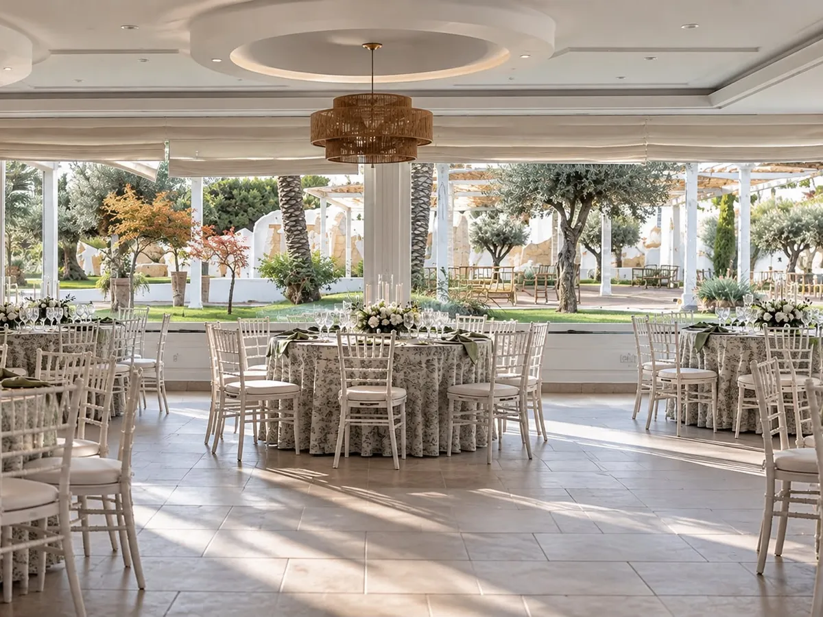 Sala interna allestita con tavoli rotondi e sedie bianche, lampadari in fibra naturale e vetrate sul giardino